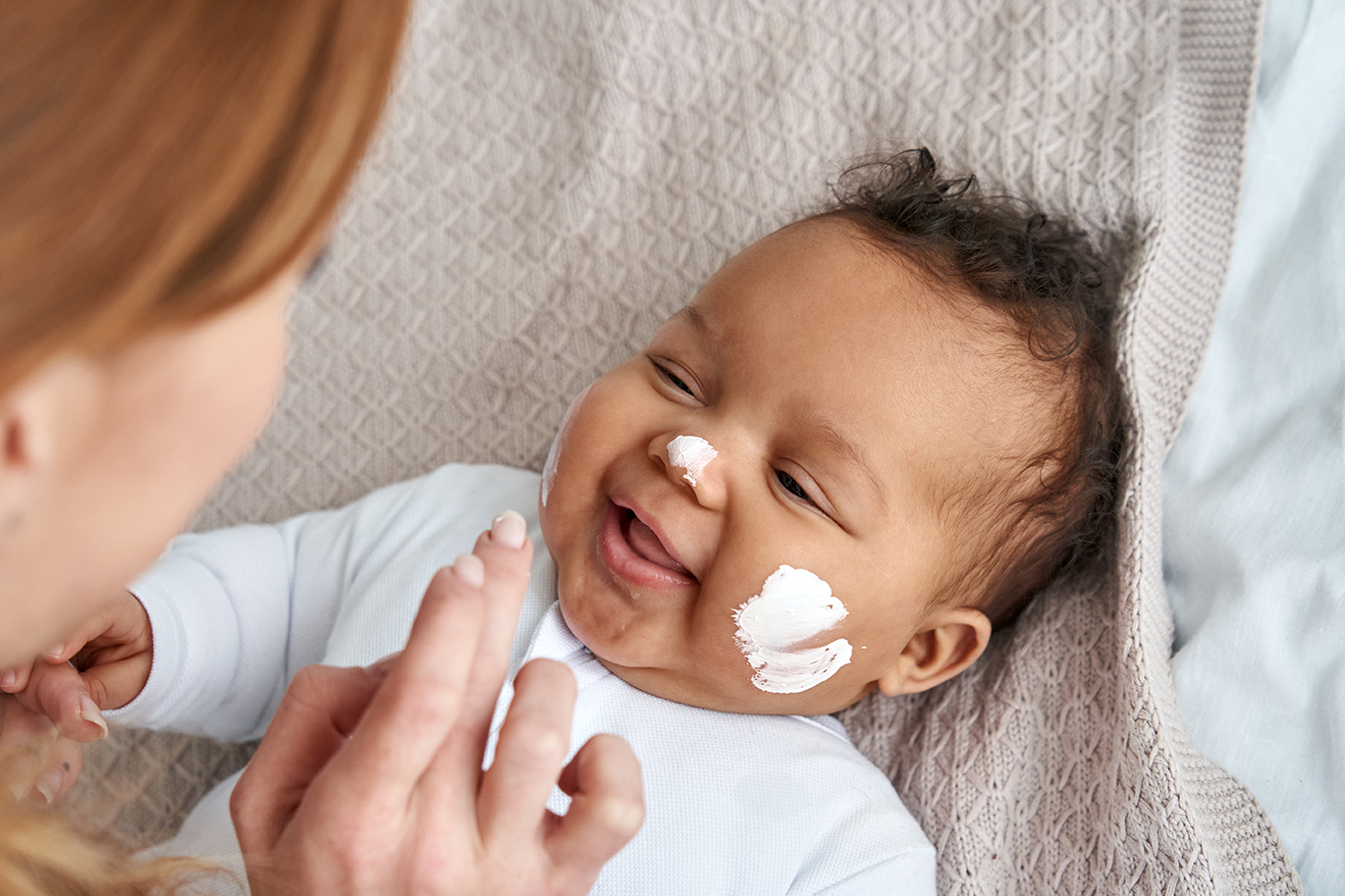 Caucasian mother applying skin care cream on cute funny african baby face.